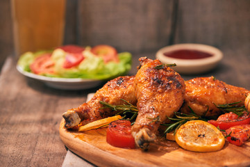 Grilled chicken legs roasted on the grill on wooden chopping board with tomato sauce in a bowl, fresh tomatoes and lettuce leaves, bitter pepper, glass mug of beer