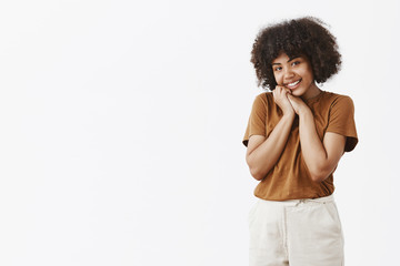 Cute and touched silly African American woman with curly hair in brown t-shirt tilting head and leaning it on hands smiling with charmed and pleased expression looking with affection at camera