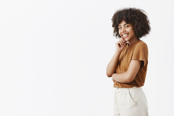 Intrigued flirty feminine dark-skinned female student with afro hairstyle in brown t-shirt standing in profile, turning at camera with cute smile holding hand on chin gazing with interest and desire