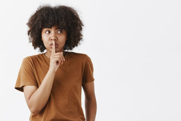 Fototapeta na wymiar Indoor shot of worried good-looking teenage african american female student with afro hairstyle saying shh while showing shush gesture feeling nervous someone know her secret or spreading rumor