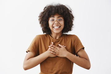 African American girl appreciate friend support being touched with kind words holding hands on heart smiling broadly with grateful happy expression standing pleased and joyful over gray background