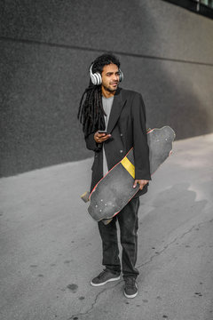 Modern urban young dreadlocks hipster man holding his skate on street and listening music. Standing against black wall with headphones on his head.