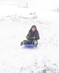 Fototapeta na wymiar Young girl slides down a hill on a toboggan on a foggy day in the snow
