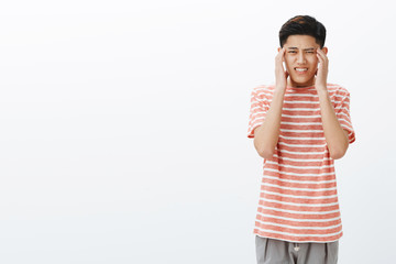 Young uneasy asian guy in striped t-shirt feeling pressured and tired holding hands on temples, suffering headache or migraine, clenching teeth from pain and intense thinking over grey wall