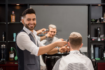 Smiling barber trimming client's stylish haircut and looking at camera.