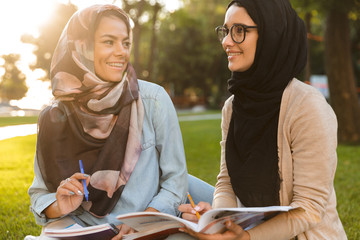 Happy young arabian women students writing in copybooks in park outdoors.