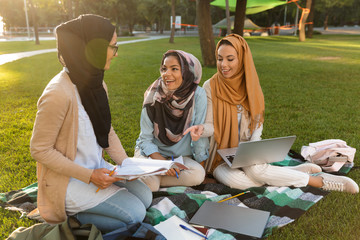 Women students using laptop computer and holding books in park.
