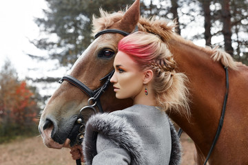 Fototapeta premium Woman on a horse in the fall. Creative bright pink makeup on the girl face, hair coloring. Portrait of a girl with a horse. Horseback riding in the autumn forest. Autumn clothes