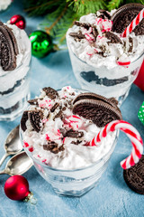 Christmas dessert, peppermint chocolate trifle in portion glasses with candy cane sweets and chocolate cookies, on light blue concrete table with xmas tree and decoration, copy space