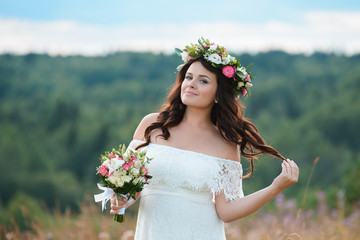 Naklejka premium Beautiful brunette girl with a bouquet and a wreath of flowers. Young woman with long curly hair in a white lace dress. Summer female portrait