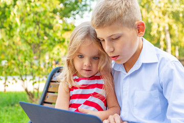 Smiling brother and sister sitting on bench in park and playing on laptop