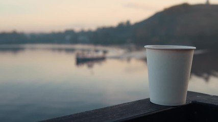 A white paper cup with hot coffee or tea stands on the railing against the backdrop of a calm river at sunset. - Powered by Adobe