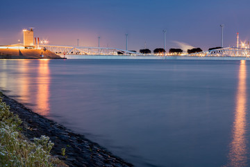 Closed and submersed Maeslant storm surge barrier at night. Dutch delta works, largest flood protection project in the world. Light reflection on water, flowers on the dyke and windmills, factories 