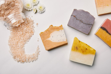 top view of various handmade soap pieces with sea salt and flowers on white marble surface