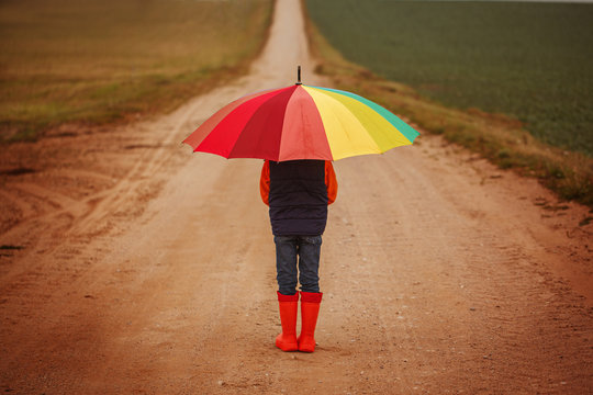 Child In Orange Rubber Boots Holding Colorful Umbrella Under Rain In Autumn. Back View