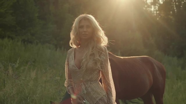 Portrait of a beautiful blonde model in open field with horse in summer, sunset light. Girl in dress posing outdoor, brown horse on a background.