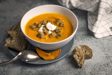 Close up of served fresh hokkaido pumpkin creamy soup with pieces of bread, baked pumkpin, potatoes and goat cheese on rustic background