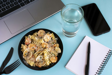 Seafood pasta business lunch at workplace. Open lunch box, open laptop, knife and fork, smartphone, glass of water, notebook, pen on blue background.