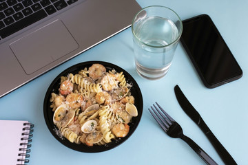 Seafood pasta business lunch at workplace. Open lunch box, open laptop, knife and fork, smartphone, glass of water, notebook on blue background.