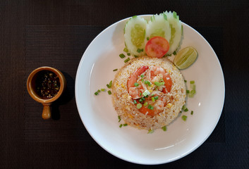 Thai style shrimps fried rice garnished with sliced cucumber, tomato,lime and onion leaves served on the white plate with fish sauce in small bowl putted on the table cloth.
