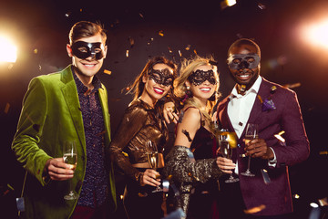 glamorous multiethnic friends in carnival masks holding champagne glasses and celebrating new year...