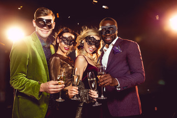 luxury multiethnic friends in carnival masks celebrating new year with champagne glasses