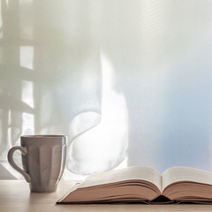 Photo of a desk with an open book and a gray cup with tea on a background of sun-lit curtains. Copy space