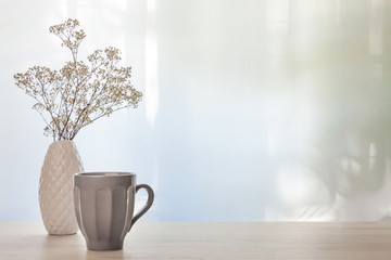 A photo of a desk with a white vase with dried white flowers and a gray cup with coffee on a...