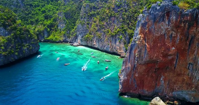 Multiple Boats in Loh Samah Bay, Thailand - Tropical Cove Surrounded by Lush Island Cliff Face With Turquoise Blue Water and Visible Coastal Reef - Traversing Overhead View