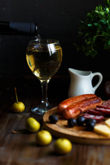 Wine pours into a glass goblet. On the table, meat appetizer, fried sausages, salami, cheese, olives.