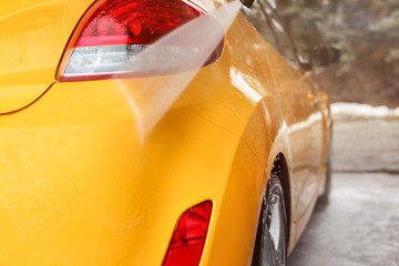 Yellow car rear light and bumper washed in self serve carwash, water jet spraying on gloss surface.