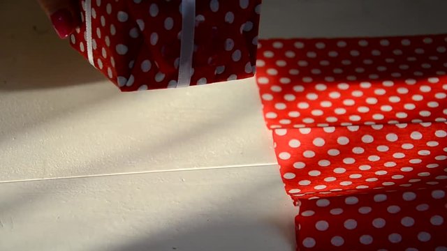 Packing presents for holiday, top view. Open a pesent. Unrecognizable woman with scissors cut ribbon and red wrapping paper in white peas for wrapping the gift box
