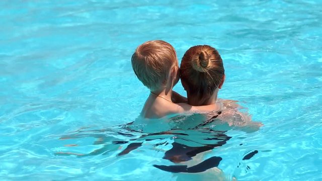 Happy young mother and her little son having fun together in an outdoor swimming pool, they laugh while swimming in the water. The kid holds on to the neck of his mother while swimming in the pool.