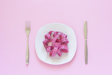 Pink measuring tape lying on plate in the form of spaghetti, knife and fork on pink background
