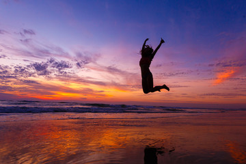 Silhouette of happy joyful woman jumping and having fun on the beach against sunset. The concept of freedom and relaxation. Reflection of the amazing sunset sky. The sky is in incredible clouds.