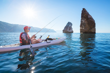 A man fishing on a kayak boat in the sea near the rocks at the shore of island mountain. Young fisherman kayaking. - obrazy, fototapety, plakaty