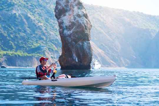 A man fishing on a kayak boat in the sea near the rocks at the shore of island mountain. Young fisherman kayaking.