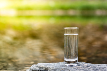 Clear Water In Clear Glass On Background Of Mountain River. Sunny Day Or Early Morning. Concept Of Healthy Food And Environmentally Friendly Natural And Crystal Clear Mineral Water.