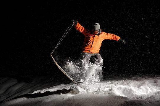Active snowboarder in orange sportswear and mask jumping on a snowy hill at night