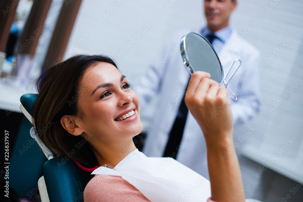 Wall mural Now my smile is perfect! Beautiful young woman looking at mirror with smile in dentist’s office