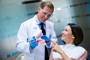 We fixed this tooth! Dentist explaining his patient about teeth and showing her plastic jaws in dentist’s office