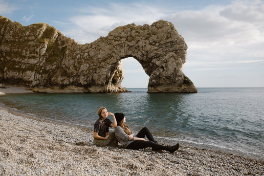 Two Woman Sitting On A Beach Looking At The Durdle Door Sea Cliff Arch In England