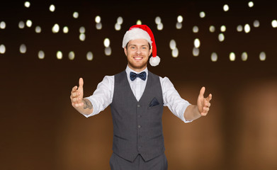 winter holidays, christmas and people concept - happy man in santa hat holding something imaginary over garland lights background
