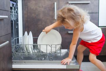 Cute blonde toddler girl helping in the kitchen taking plates out of dish washing machine
