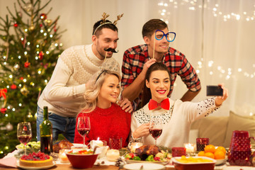 holidays and celebration concept - happy friends with party props taking selfie by smartphone at home christmas dinner