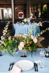 Festive table decorated with white plates and flowers on a hight silver vase on blue tablecloth