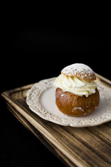 Tasty small sweet cake with whipped cream and powdered sugar on top on a wooden tray, black background.