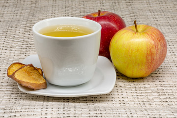 A cup of apple tea with two apples and dehydrated slices of the fruit with a patterned table cloth in the background