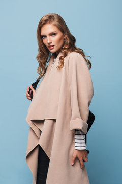 Young attractive woman with wavy hair in beige coat with black backpack thoughtfully looking in camera over blue background isolated