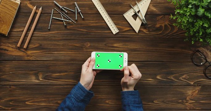 Top view on the white smartphone lying horizontally on the wooden table with office supplies and nails, female hands scrolling and tapping on the green screen. Chroma key. Tracking motion.